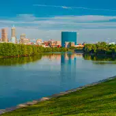 Panoramic view of Indianapolis skyline with modern buildings, lush green trees, and clear blue water reflecting the cityscape under a bright sky.