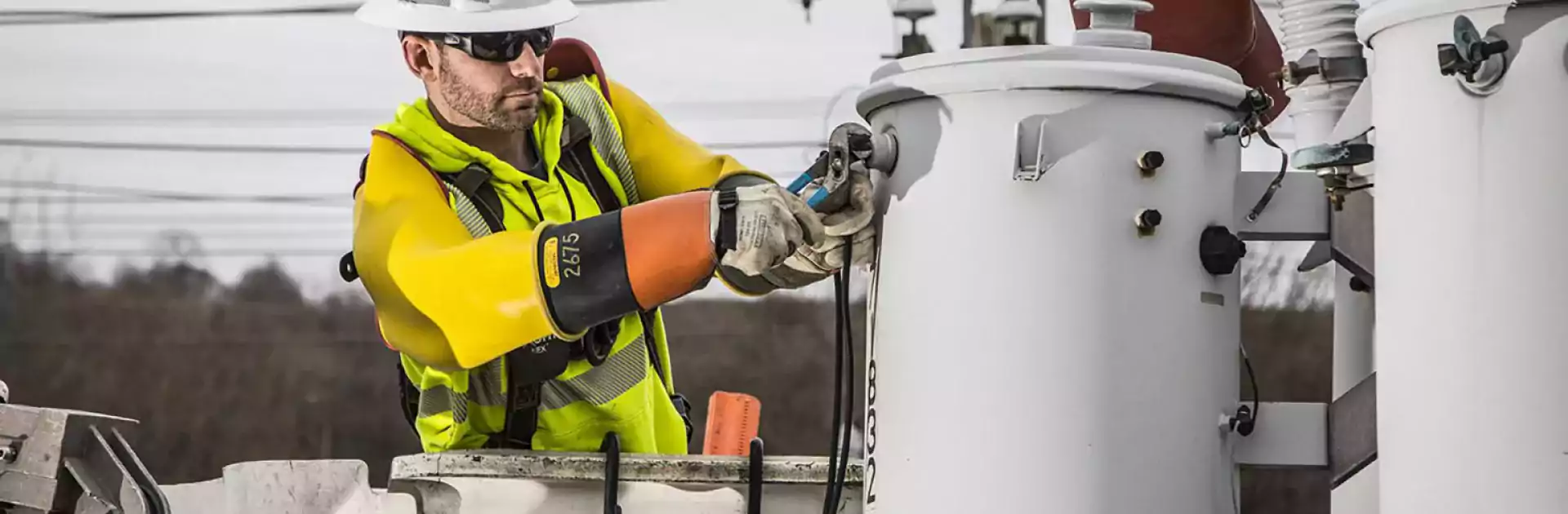 Utility worker wearing safety gear and insulated gloves repairs electrical equipment on a power pole using tools, standing in a bucket truck on an overcast day.