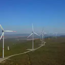 A row of wind turbines stands on a green landscape under a clear blue sky, generating renewable energy in a rural area.