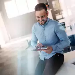 Smiling man in a blue shirt sits on a desk in a modern office, using a digital tablet. Bright natural light comes through large windows, creating a professional and relaxed atmosphere.