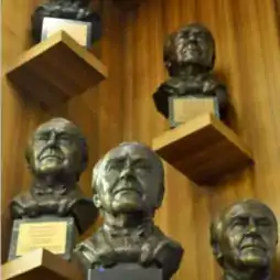 Five bronze bust sculptures of men displayed on wooden shelves against a wood-paneled wall, each with a plaque at the base.