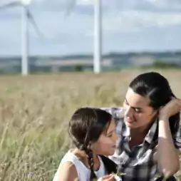 Mother and daughter sitting in a grassy field with wind turbines in the background on a sunny day, highlighting a connection with nature and renewable energy.