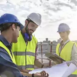 Three construction workers wearing safety vests and helmets are discussing blueprints at a construction site outdoors. One worker holds a clipboard, while the others review the plans together under a partly cloudy sky.