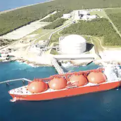 Aerial view of a large red LNG tanker docked at a coastal terminal with spherical storage tanks and industrial buildings surrounded by greenery.