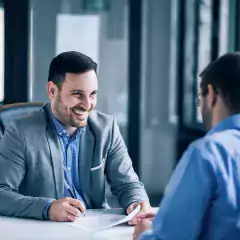 Two men in business attire sitting at a desk in a modern office, smiling and discussing documents together in a professional meeting.