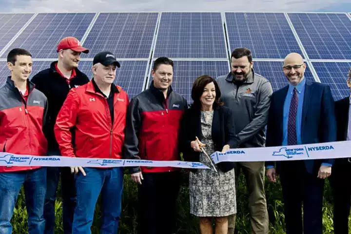 A group of people standing in front of solar panels at a ribbon-cutting ceremony for a solar energy project. The group includes men and women dressed in business and casual attire, holding a ribbon marked with NYSERDA and New York State logos.
