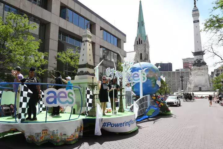 A colorful parade float with people waving, featuring wind turbines, a globe, and the AES logo, moves through a city street with historic buildings and a monument in the background. The float promotes renewable energy and community involvement.