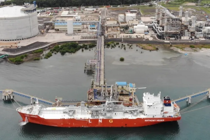 Aerial view of a liquefied natural gas (LNG) terminal with a large red and white LNG tanker docked at the pier, pipelines connecting the ship to storage tanks, and an industrial facility with storage tanks and processing units in the background.