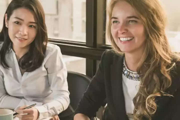 Two women in business attire sitting at a table in a bright office, one smiling and the other holding a pen, engaged in a meeting or discussion.