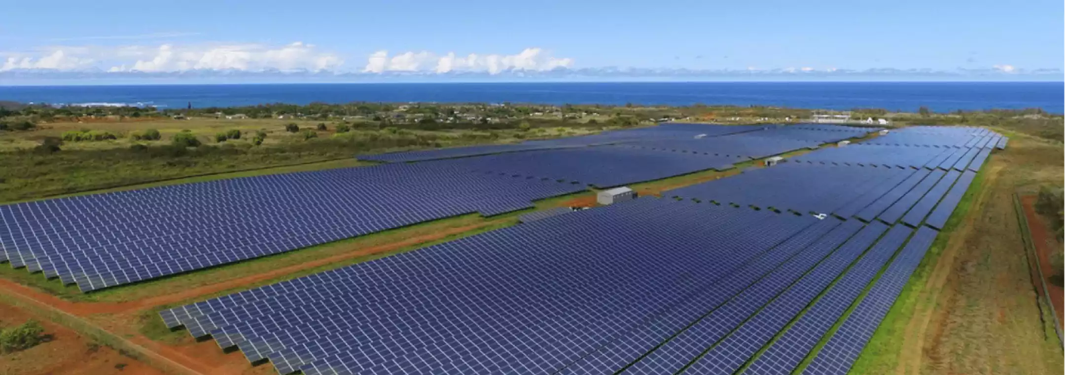A large solar farm with rows of solar panels stretches across a green landscape near the coastline under a clear blue sky.
