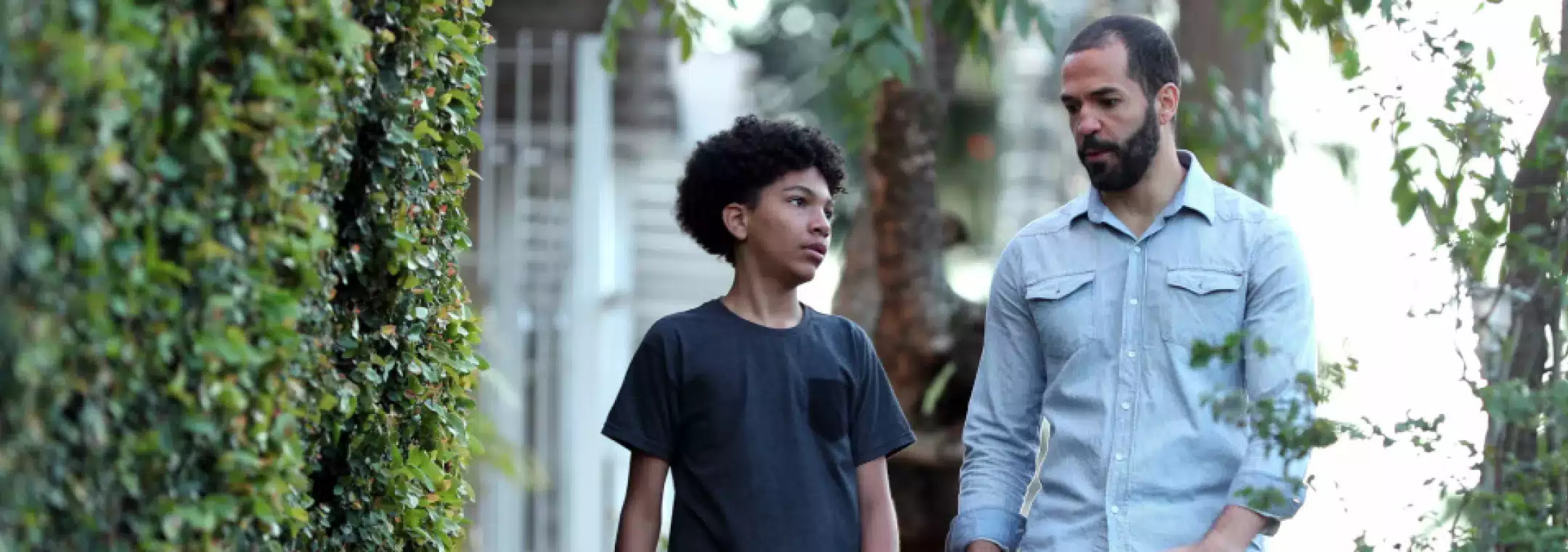 An adult man and a young boy are walking together outdoors along a path lined with green bushes, engaged in a serious conversation.