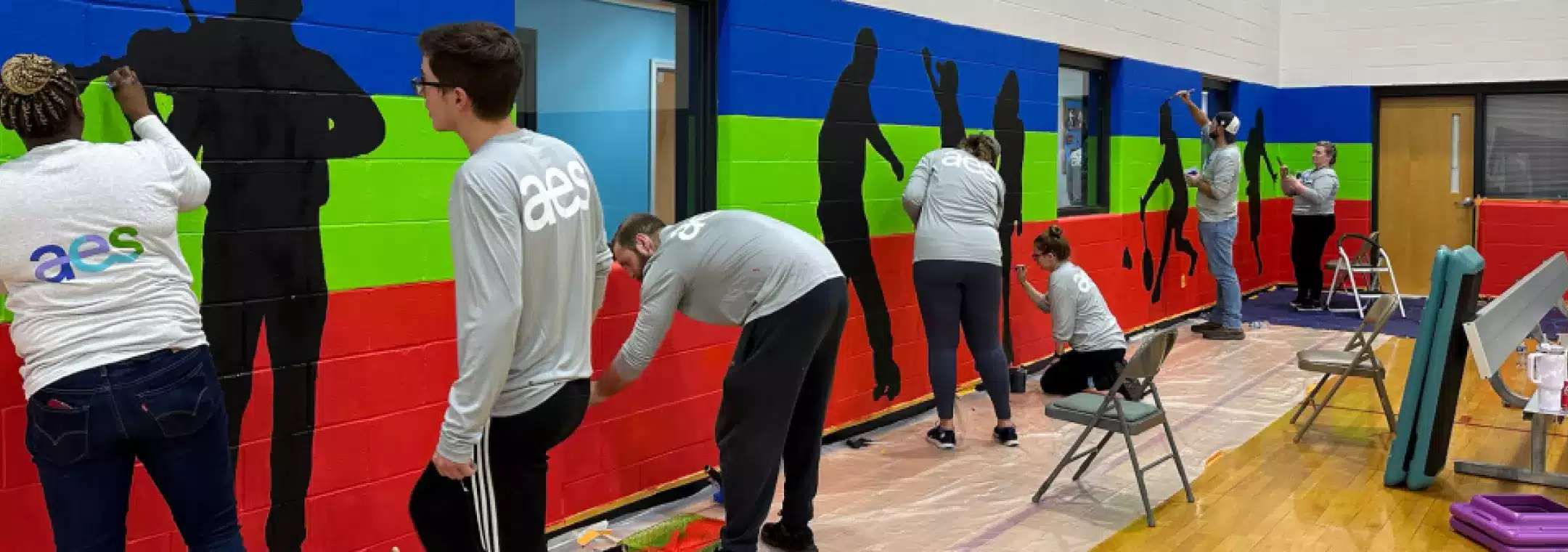 A group of people wearing matching AES shirts paint colorful silhouettes on a gymnasium wall. The mural features black human figures against blue, green, and red horizontal stripes. Ladders, chairs, and painting supplies are visible on the gym floor.