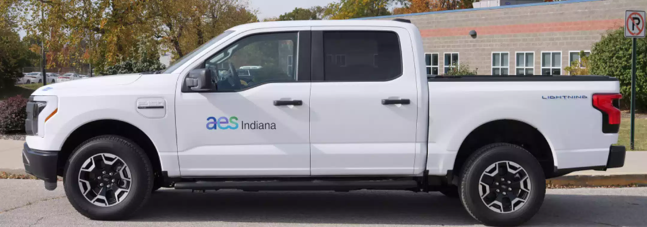 A white Ford F-150 Lightning pickup truck with AES Indiana branding is parked on a street near a building with large windows and autumn trees in the background.