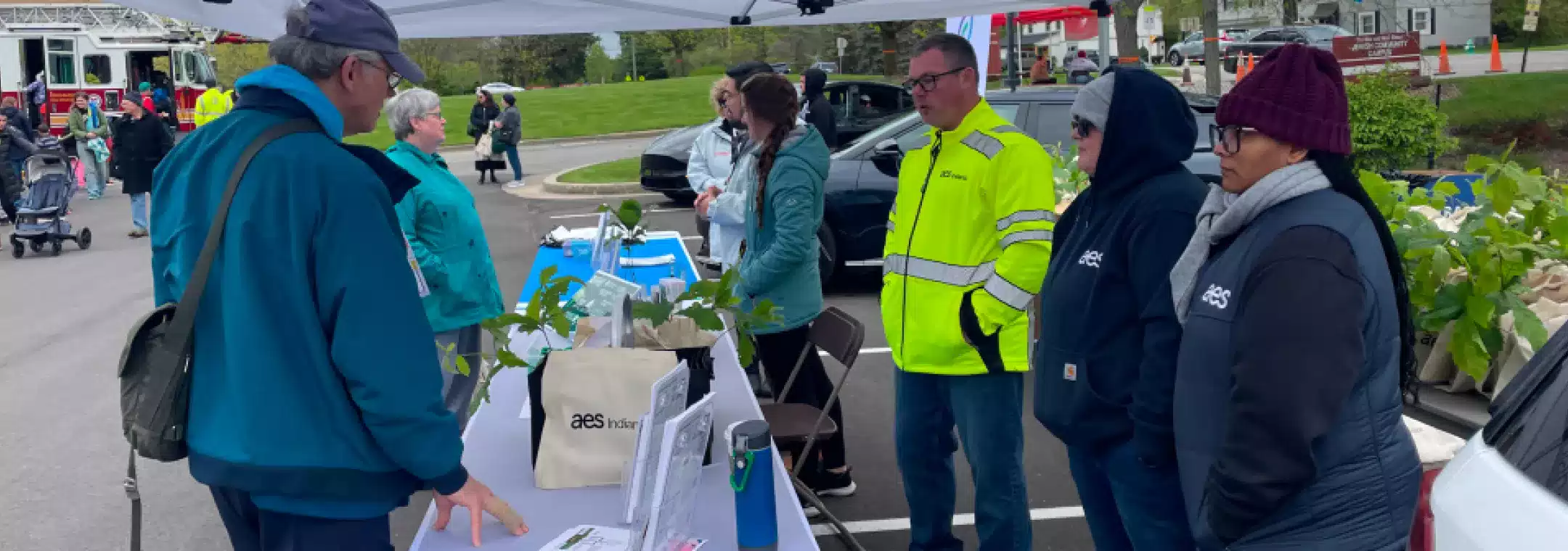 People gather at an outdoor community event booth with informational materials and plants on the table. Several individuals in jackets and hats interact under a canopy, while others walk in the background near a fire truck and green lawn.