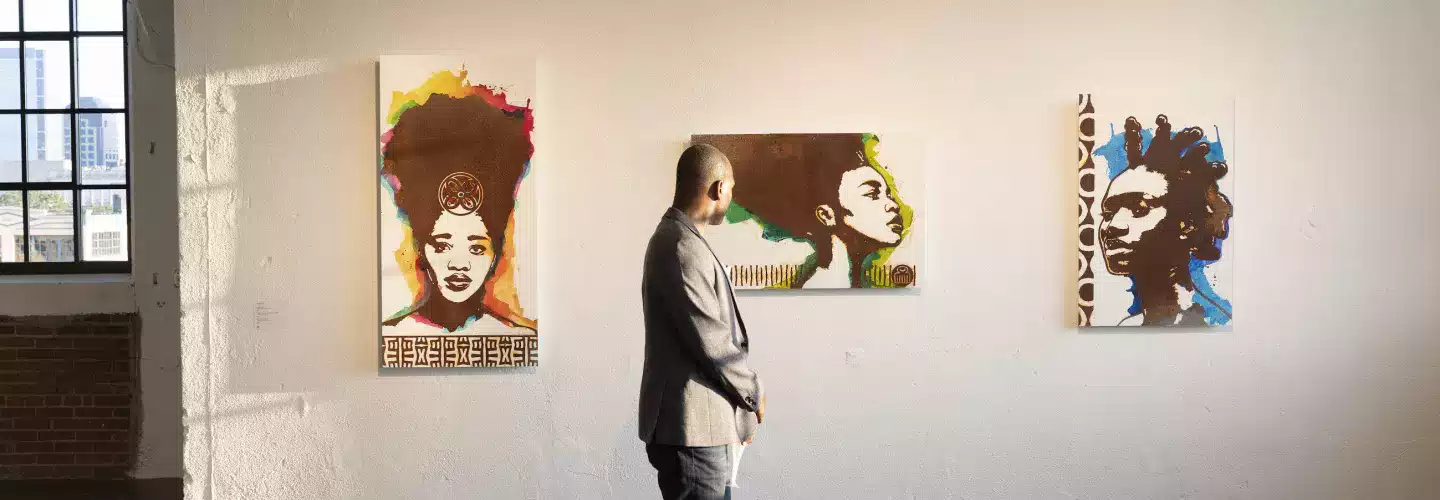 A man in a gray blazer stands in an art gallery, observing three colorful portraits of women with natural hairstyles displayed on a white wall. Sunlight streams in from a window, creating a bright and inviting atmosphere.