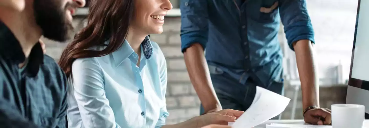 Four young professionals collaborate at a desk in a modern office, smiling and looking at a computer screen together. Papers, a keyboard, and a coffee mug are on the desk, with a whiteboard and monitor in the background.