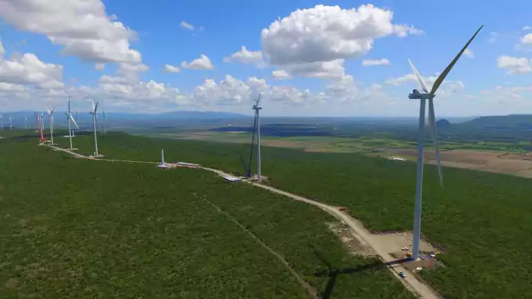 Aerial view of a wind farm with multiple large wind turbines spread across a green landscape under a blue sky with scattered clouds. Some turbines are under construction with cranes and vehicles visible along a dirt road.