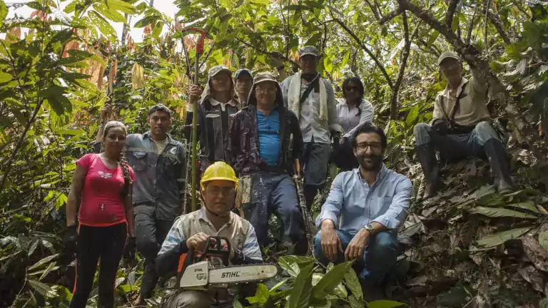 A group of people, including men and women, pose together in a lush forest or jungle setting. Some wear work clothes and hats, and one person holds a chainsaw, suggesting they are involved in forestry or agricultural work.