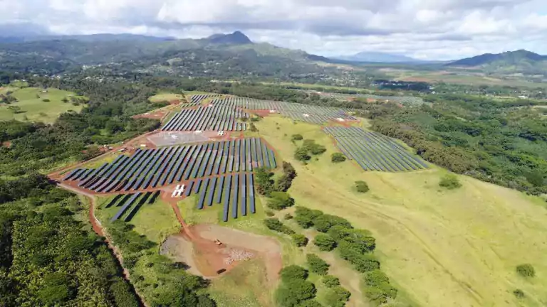 Aerial view of a large solar farm with rows of solar panels installed on green fields, surrounded by trees and hills under a partly cloudy sky.