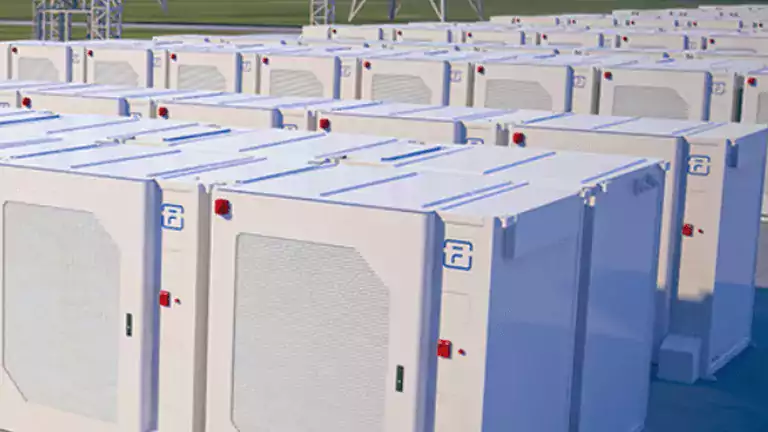 Rows of large white battery storage units are installed outdoors near high-voltage power lines and electrical towers, with a green landscape and hills in the background.