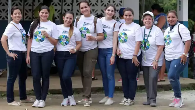 A group of eight women standing outdoors, smiling and wearing matching white t-shirts with a colorful circular logo, dark pants, and casual shoes. The background shows a building with a patterned wall and greenery.
