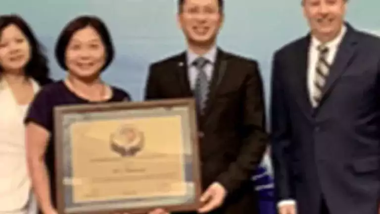 Four people stand together indoors, with one woman holding a framed certificate. All are dressed formally and smiling, posing against a light blue background.