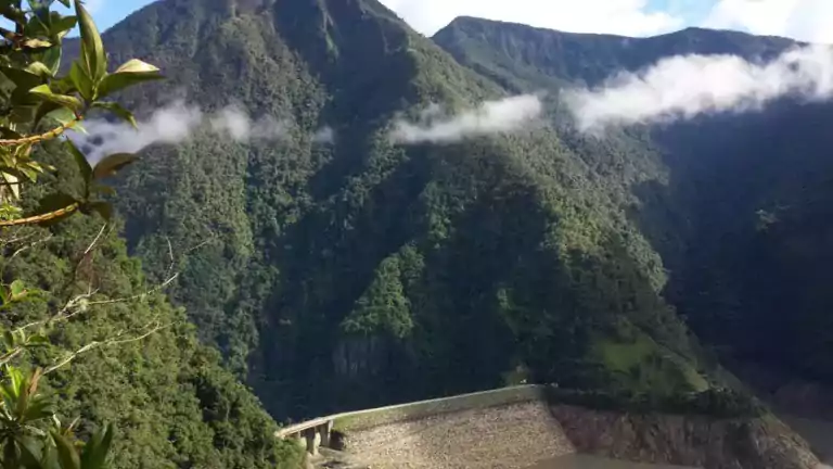 A large dam at the base of lush, green mountains with low clouds drifting across the slopes. The dam holds back a reservoir, and dense forest covers the landscape. Sunlight highlights the mountainous terrain and vegetation.