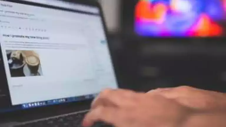 Close-up of hands typing on a laptop keyboard, with a blog post editor open on the screen. A blurred television with colorful lights is visible in the background.