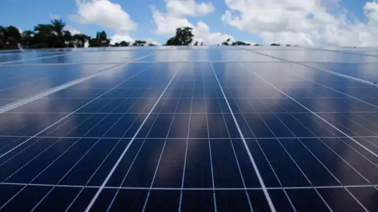 Close-up view of solar panels reflecting the sky with scattered clouds, surrounded by trees in the background.