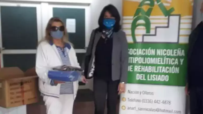 Three people wearing face masks stand indoors next to a banner for the Asociación Nicoleña de Rehabilitación del Lisiado. One person holds a blue plastic container. An open cardboard box is on a nearby table.
