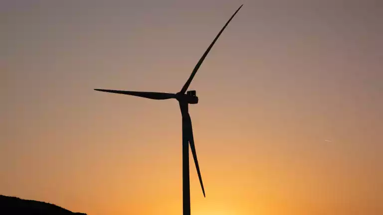 A wind turbine stands silhouetted against an orange sunset sky, with distant hills visible at the bottom of the image.