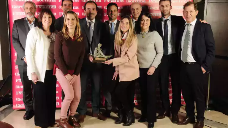 A group of eleven people, both men and women, stand together smiling and posing for a photo in front of a red Fortuna backdrop. Two people in the front center are holding a trophy. The group appears to be celebrating an award or achievement.
