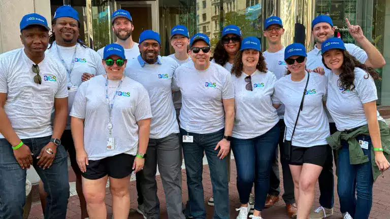 A diverse group of people wearing matching gray AES Indiana t-shirts and blue hats pose together outside an office building, smiling and standing close as a team.