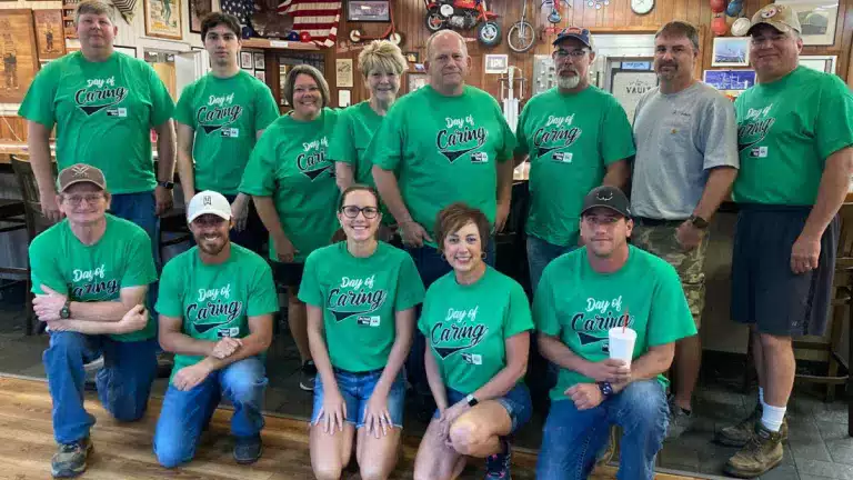 Group of volunteers wearing matching green 'Day of Caring' shirts pose together indoors, smiling at the camera. The setting features wooden walls, framed pictures, and American-themed decor, indicating a community or volunteer event.