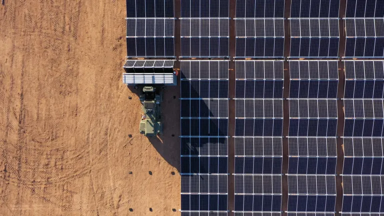 Aerial view of a solar panel installation site showing a vehicle and worker placing solar panels on a large array in a dry, sandy area.