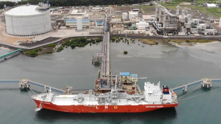 Aerial view of a liquefied natural gas (LNG) terminal with a large red and white LNG tanker docked at the pier, pipelines connecting the ship to storage tanks, and an industrial facility with storage tanks and processing units in the background.