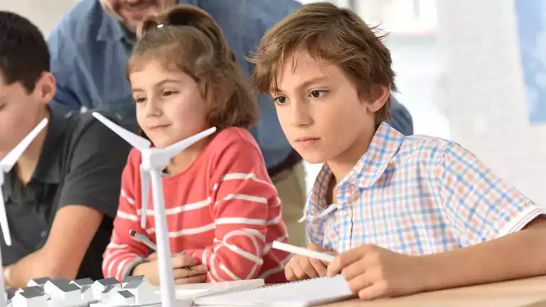 Children in a classroom closely observe a model of wind turbines and houses on a table, guided by an adult, illustrating a renewable energy or science lesson.