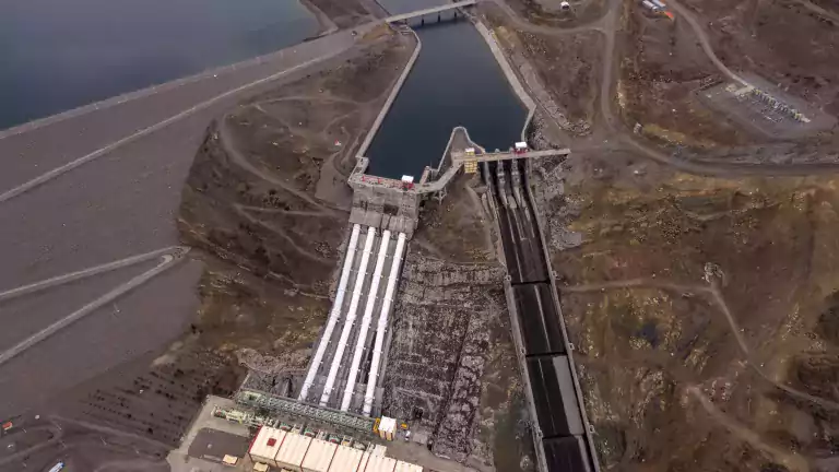 Aerial view of a large hydroelectric dam with a reservoir, spillways, and surrounding rocky landscape. The image shows water intake structures, power generation buildings, and access roads leading to the facility.