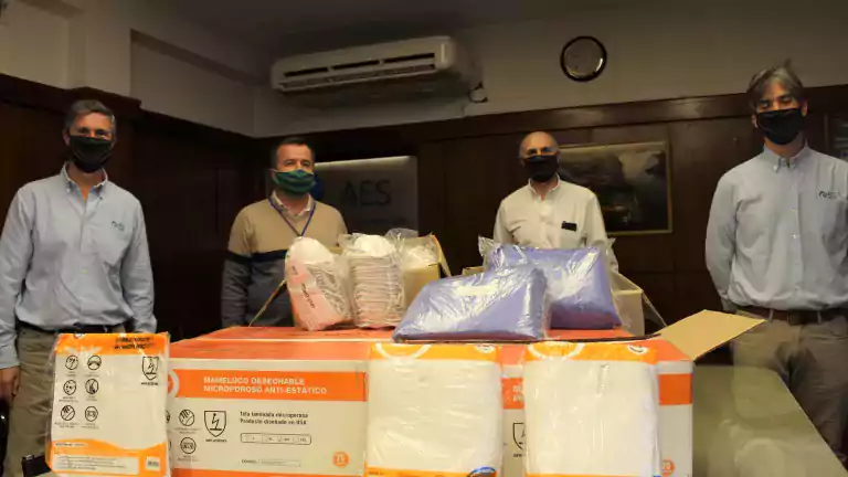 Four men wearing face masks stand behind a table with boxes and packages of disposable protective coveralls and personal protective equipment in an office setting.