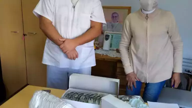 A healthcare worker and a woman wearing face masks and protective gear stand behind a table with boxes of medical supplies, including face shields and masks, in an office setting.