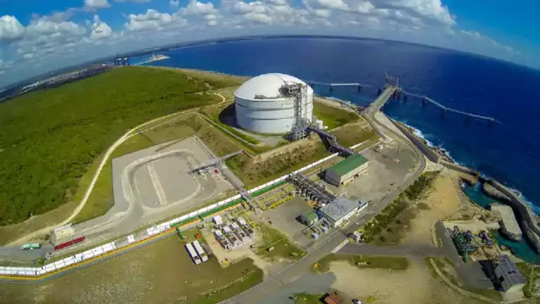 Aerial view of a coastal industrial facility with a large white storage tank, several buildings, roads, and a pier extending into the blue ocean, surrounded by green forest and partly cloudy sky.