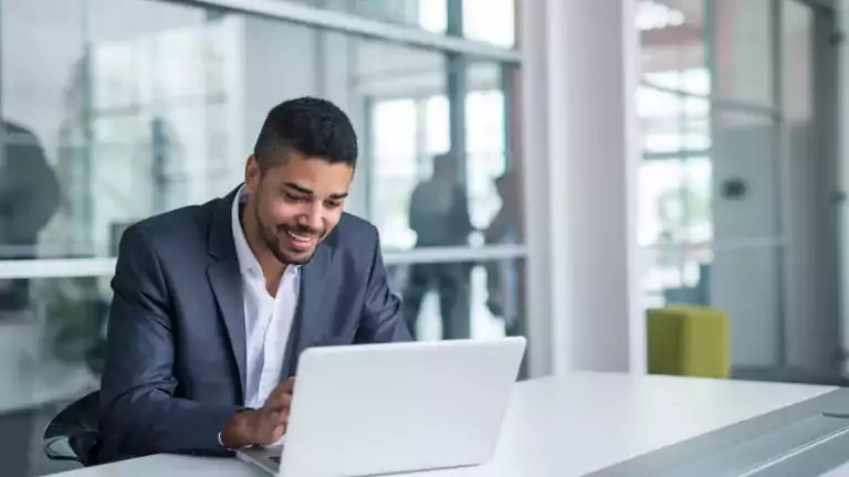 Smiling businessman in a suit working on a laptop at a modern office desk with glass walls and natural light.