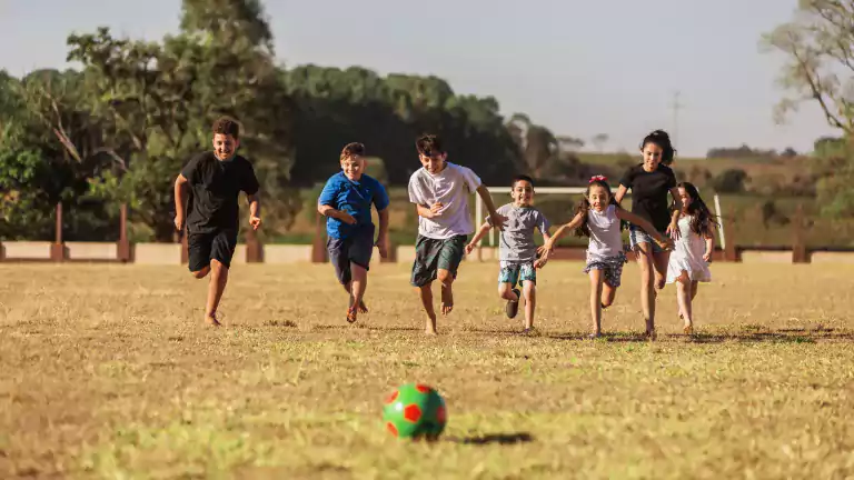 A group of seven children run barefoot on a grassy field toward a colorful soccer ball, smiling and playing together on a sunny day with trees and a soccer goal in the background.