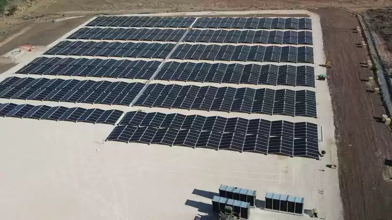 Aerial view of a large solar farm with multiple rows of solar panels installed on a cleared, flat area surrounded by trees and dirt roads. A vehicle and several containers are visible near the panels.