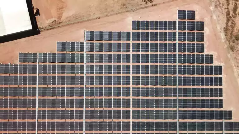 Aerial view of a large solar panel array installed on a flat, reddish dirt ground with sparse trees and industrial equipment nearby, demonstrating renewable energy generation in a rural area.