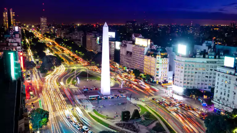 Night view of Buenos Aires city with the illuminated Obelisk in the center, surrounded by busy streets with light trails from traffic and tall buildings lit up against a dark sky.