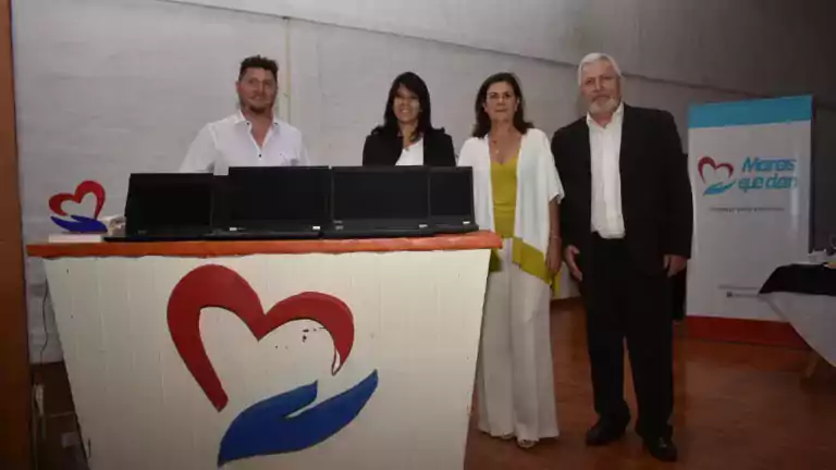 Four people stand behind a table with laptops and a heart and hand logo in a room with wooden floors. A banner in the background reads 'Manos que dan' and indicates a socio-educational project.