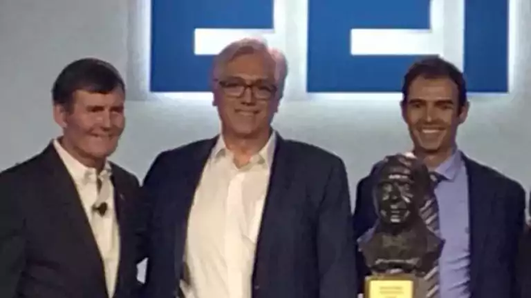 Four people in business attire stand together on stage, smiling and posing with a bronze bust trophy. A geometric design and blue letters are visible in the background.