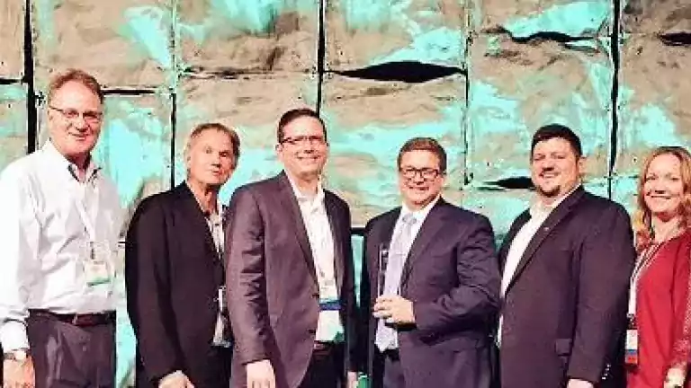 A group of eleven professionally dressed people stand side by side, smiling at the camera in front of a textured, illuminated conference backdrop.