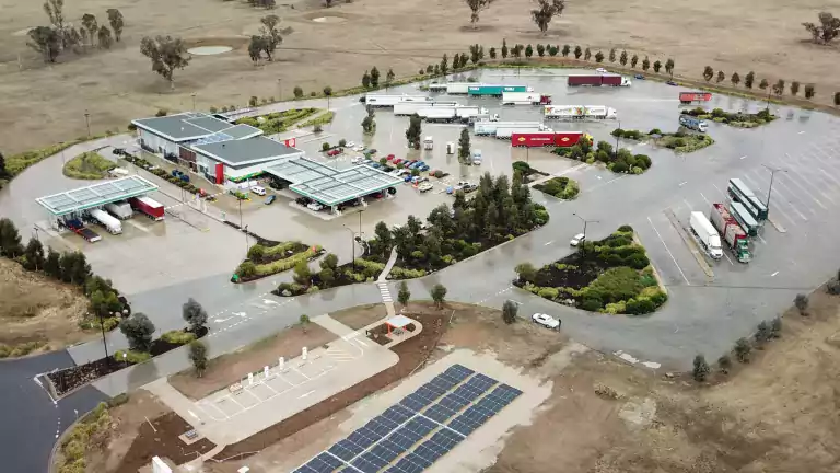 Aerial view of a rural truck stop with fuel stations, parking areas for trucks and cars, a main building, and a solar panel array in the foreground. The complex is surrounded by dry grassland and scattered trees, with hills visible in the background.